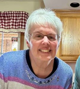 An older woman with short white hair and glasses smiles indoors, wearing a blue sweater with sequins. Wooden cabinets and a checkered curtain are in the background.