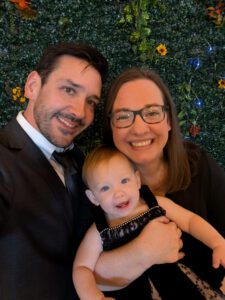 A man, woman, and baby smile for a selfie in front of a green leafy background decorated with small flowers.