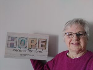 An older woman wearing glasses and a pink sweater holds a sign that reads, "HOPE anchors the soul. Hebrews 6:19" against a plain wall.
