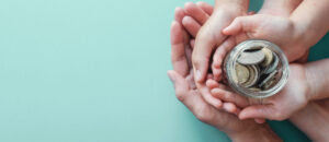 Adult and child hands hold a glass jar filled with coins against a light blue background.