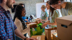 A group of people stand around a table sorting and handing out boxes and bags, appearing to participate in a community volunteer event indoors.