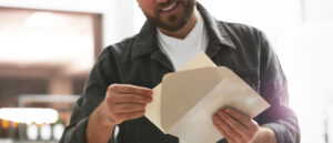 A man wearing a white t-shirt and dark jacket smiles while opening a beige envelope and holding a card indoors.