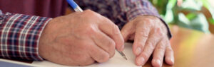 Close-up of an elderly person’s hands writing a letter with a blue pen at a wooden table, with papers and a plant visible in the background.