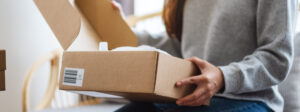 A woman in a gray sweater opens a cardboard box while sitting indoors. The box has a barcode label and packing material inside.