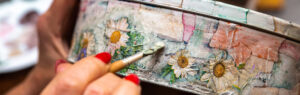 Close-up of hands with red nail polish painting white daisies on a round tin container using a small brush.