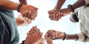 Group of people standing in a circle, holding hands with fingers interlocked, photographed from below against a cloudy sky.