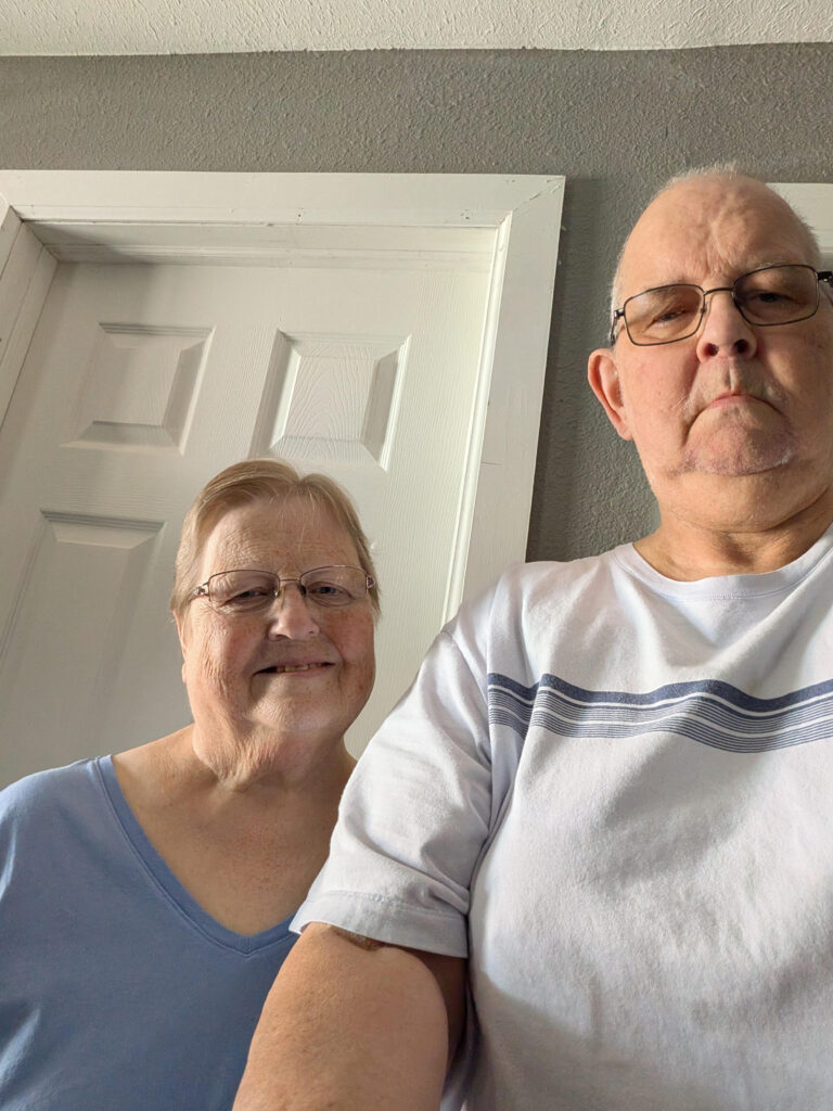 Two older adults stand indoors in front of a white door, both wearing glasses and casual shirts, looking at the camera.
