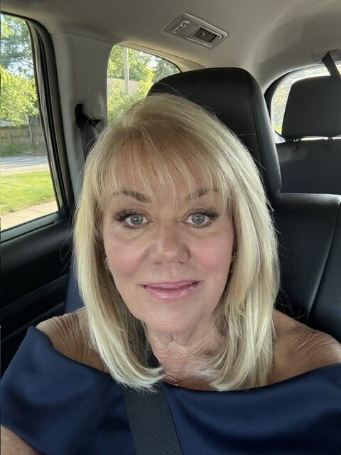 A woman with blonde hair and an off-shoulder top sits in the front seat of a car, wearing a seatbelt, facing the camera.