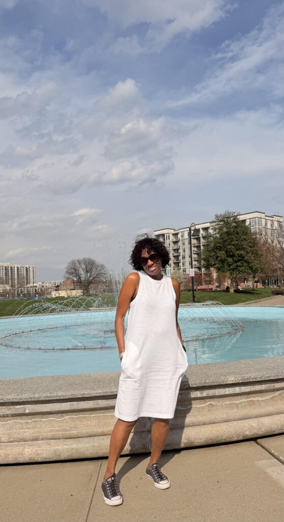 A person in a sleeveless white dress stands in front of a large fountain, with modern buildings and trees in the background on a partly cloudy day.