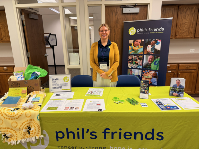 A woman stands behind a Phil’s Friends booth with pamphlets, gift bags, and promotional items on display in a room with wooden cabinets.