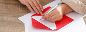 A person writing on a note card placed inside a red envelope on a wooden desk.